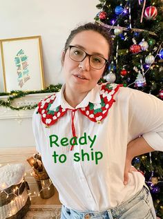 a woman standing in front of a christmas tree wearing a shirt that says ready to ship