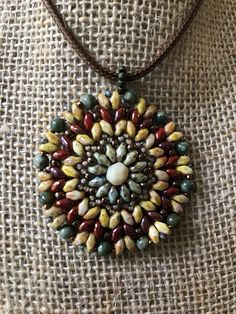 This colorful hand-beaded Mandala Necklace is made using many different style beads, and as you can see, very colorful!  It is warm colors, which include greens, browns, deep red, and brass colored beads. These include Super Duo beads, as well as assorted round beads in various sizes, and a large cut bead as the center piece. It is quite flexible, as shown in the one picture, but lays flat against your body. It measures 1-3/4 " inches in diameter, not including the beaded bale. It also includes Bohemian Round Brown Beads, Bohemian Brown Beaded Necklace, Bohemian Brown Beaded Round Necklaces, Brown Beaded Necklace With Colorful Oval Beads, Bohemian Beaded Necklaces With Faceted Beads, Bohemian Brown Beaded Necklaces With Oval Beads, Handmade Brown Czech Glass Beads, Earthy Multicolor Gemstone Beaded Necklaces, Artisan Brown Spacer Beads