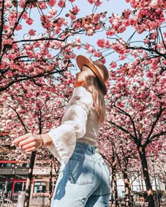 a woman wearing jeans and a hat standing in front of trees with pink flowers on them