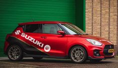 a red and black suzuki car parked in front of a green garage door with the word suzuki on it