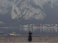 a woman standing on the side of a road next to a body of water with mountains in the background