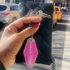 a person holding up a pink keychain on a black purse with cars in the background