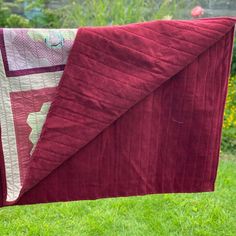 a red and white quilt hanging from a clothes line in the grass with flowers behind it