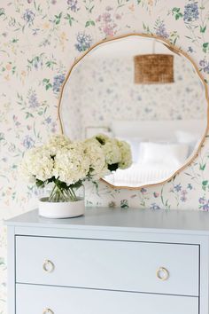 a white dresser with flowers and a round mirror on the wall above it, in front of a floral wallpapered bedroom