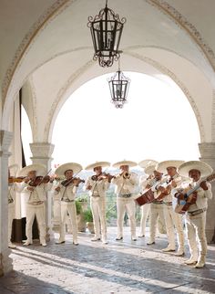 a group of men in white suits and hats are playing instruments under an archway with a chandelier hanging from the ceiling