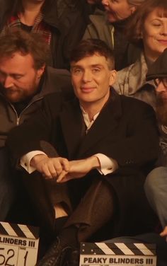 a man sitting in front of a crowd wearing a black suit and hat with his hands on his knees