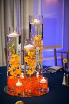 two tall clear vases with yellow flowers and candles are on a blue table cloth