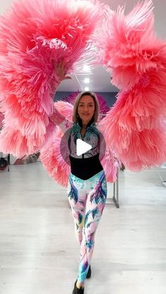 a woman is standing in front of some pink feathers on her head and holding up two large pink wings