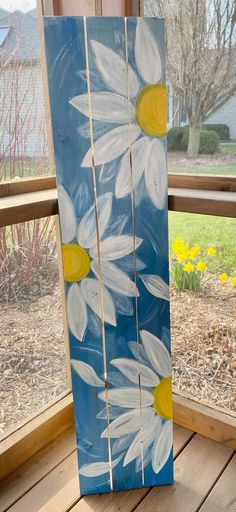 a painted wooden board with white daisies on it sitting in front of a window