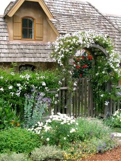 a garden with white flowers and greenery next to a wooden fence in front of a house