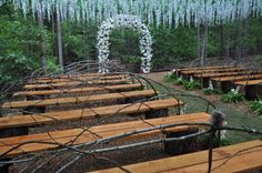 an outdoor ceremony setup with wooden benches and white flowers hanging from the trees in the background