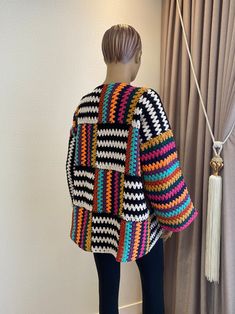 a woman standing next to a window wearing a multicolored crocheted jacket