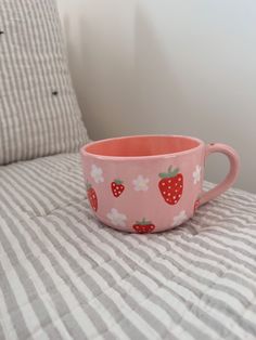 a pink cup with strawberries on it sitting on a bed next to striped pillows