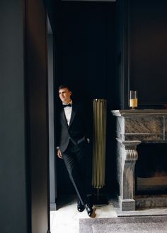 a man in a tuxedo leaning against a fireplace