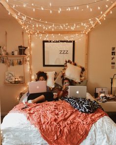 two people sitting on a bed with laptops in front of their faces and lights strung across the room