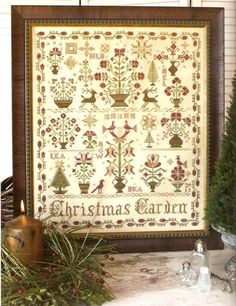 a cross - stitch christmas sample is displayed in front of a candle holder and potted plant