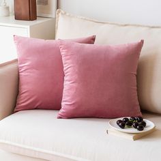 two pink pillows sitting on top of a white couch next to a plate of fruit