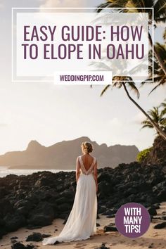 Bride in white gown stands on rocky shore at sunset with mountains and palm trees in the background. Lush Garden