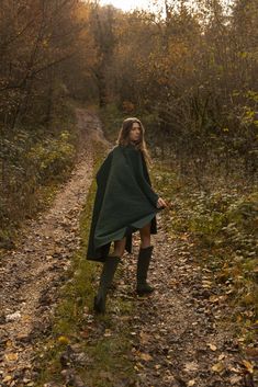 a woman standing on a dirt road in the woods wearing a green cape and boots