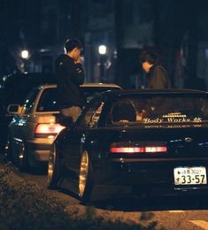 two men standing next to cars on the side of the road at night with one man talking on his cell phone