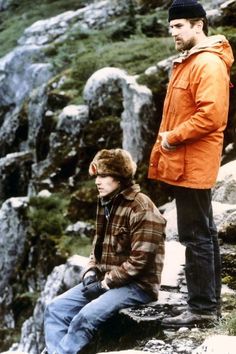 two young men sitting on rocks in the mountains, one is wearing an orange jacket