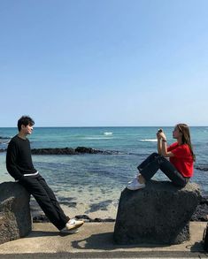 two people sitting on rocks near the ocean and one person is taking a photo with his cell phone