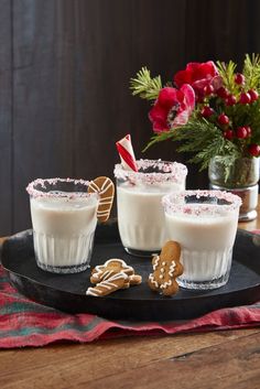 three glasses filled with milk and cookies on a black platter next to red flowers