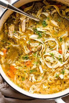 a pot filled with chicken noodle soup on top of a white cloth next to a spoon