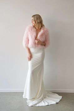 a woman in a long white dress and pink fur stoler standing against a wall