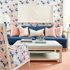 a living room with floral wallpaper and blue couches, pink pillows, and white coffee table