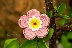 a crocheted pink flower sitting on top of a green leaf covered tree branch