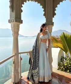 a woman in a white and blue lehenga stands on a balcony overlooking the water