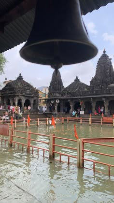 a bell hanging from the side of a building next to a body of water with people standing around it