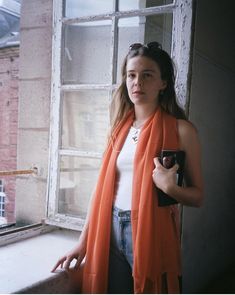 a woman standing in front of a window with an orange scarf around her neck and holding a cell phone