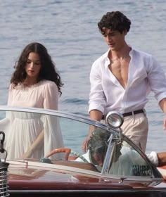 a man and woman standing next to a boat in the water with one holding on to the steering wheel