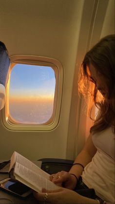 a woman sitting on an airplane reading a book and looking out the window at the sky