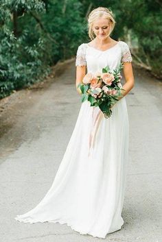 a woman in a long white dress holding a bouquet and standing on a road surrounded by trees