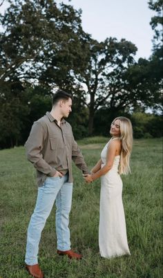 a young man and woman holding hands in the middle of a field with trees behind them