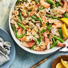 a white bowl filled with pasta, shrimp and asparagus next to lemon wedges