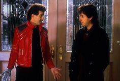 two men standing next to each other in front of a door wearing red jackets and black pants