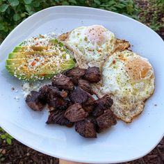 an egg, steak and avocado on a white plate in front of some bushes