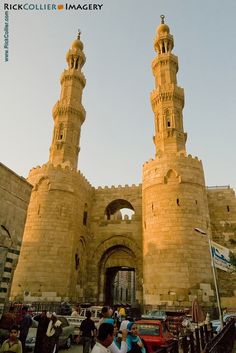 two tall towers are in the middle of an old city square with people walking around