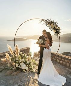 a bride and groom standing in front of an arch with flowers on it at sunset