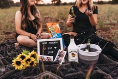 two women sitting on a blanket with drinks and snacks in their hands, one holding a beer