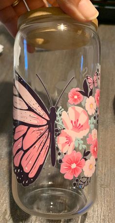 a hand is holding a glass jar with a butterfly painted on the side and pink flowers