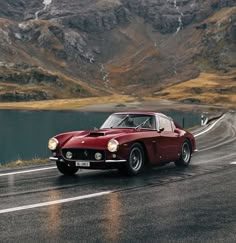 an old red sports car driving down the road in front of some mountains and water