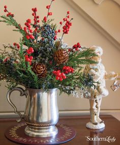 a silver mug with red berries and pine cones in it sitting on a table next to a figurine