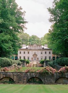 a large white house surrounded by lush green trees and flowers in front of a stone bridge