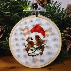 a cross stitch christmas ornament hanging from a tree with pine cones in the background