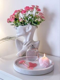 a white vase with pink roses in it and a perfume bottle on a tray next to a candle